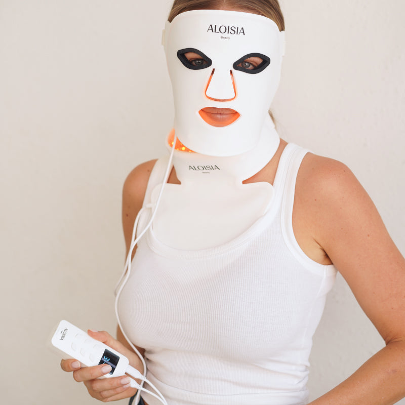 Woman wearing the REFINING LED face mask and neck panel lit red, holding the controller