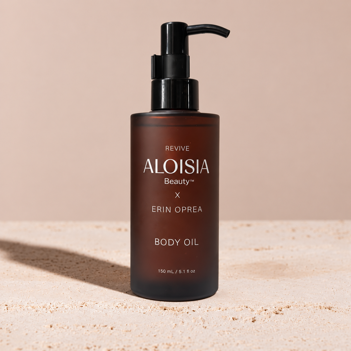 A 150 mL bottle of ALOISIA Beauty REVIVE Body Oil x ERIN OPREA in a frosted amber-brown glass bottle with a black pump dispenser, set on a sandy beige surface against a solid beige background.